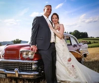 Bride and groom with a vintage car in a field – wedding photo with floral dress by hochzytfotograf.com